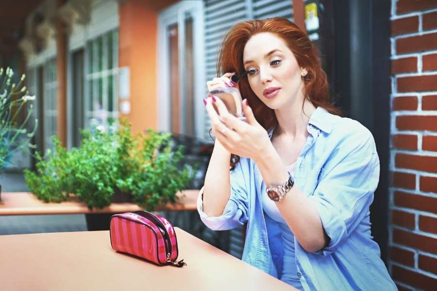 Une femme rousse assise à une terrasse applique rapidement son maquillage express avec un miroir de poche.