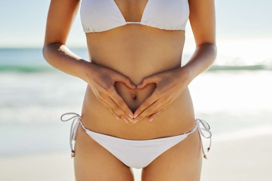Une femme en bikini blanc forme un cœur avec ses mains sur son ventre face à la mer.
